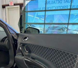 Interior view of a car door with quilted leather paneling, a metallic door handle, and speaker detailing. The window is partially open, and a sign reading "COAT DETAILING" and "PAINT PROTECTION FILM" is visible through the tinted glass.
