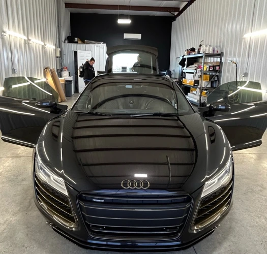 Front view of a black Audi R8 with its doors open inside a detailing garage. The vehicle’s glossy finish reflects the overhead lights, and two workers are seen in the background preparing equipment.