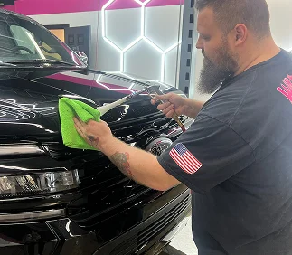 Man using a spray tool and green microfiber cloth to clean the front grille of a black SUV in a well-lit auto detailing shop. He wears a dark shirt with an American flag patch on the sleeve.