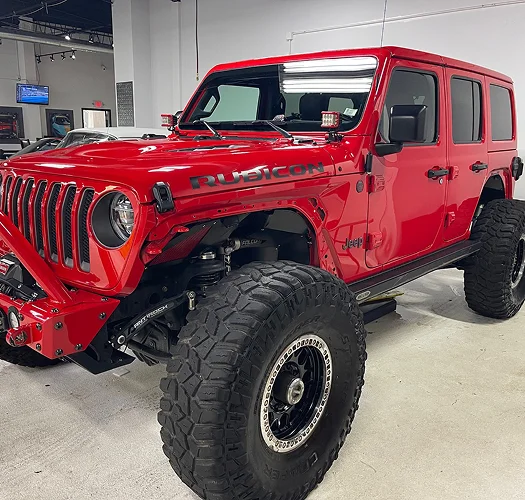 Bright red Jeep Wrangler Rubicon with large off-road tires, lifted suspension, and aftermarket front bumper parked inside a detailing or modification shop.
