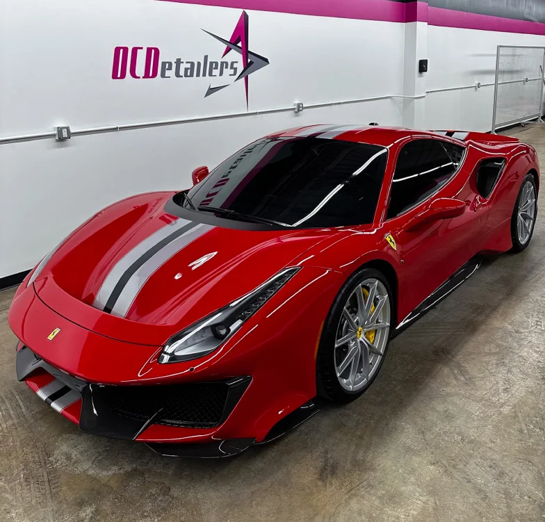 Glossy red Ferrari with dual racing stripes in black and silver parked inside a detailing shop with "OCDetailers" branding on the wall. The supercar features sleek curves, sharp headlights, and silver alloy wheels.