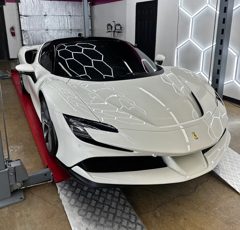 Glossy white Ferrari SF90 Stradale parked inside a detailing studio, positioned on a red and silver platform with geometric LED wall lights reflecting off its polished surface.