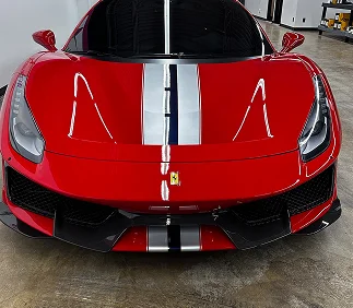 Front view of a red Ferrari with a silver and black racing stripe running down the center of the hood, parked on a polished concrete floor in a detailing shop.