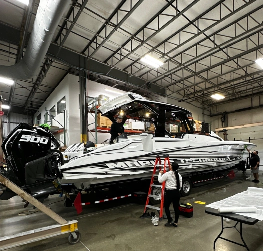Large white powerboat with “Marine Technology” graphics being worked on in an indoor facility. The boat is mounted on a trailer with workers using a ladder and equipment for detailing or maintenance. Dual black Mercury 500 outboard motors are visible at the rear.