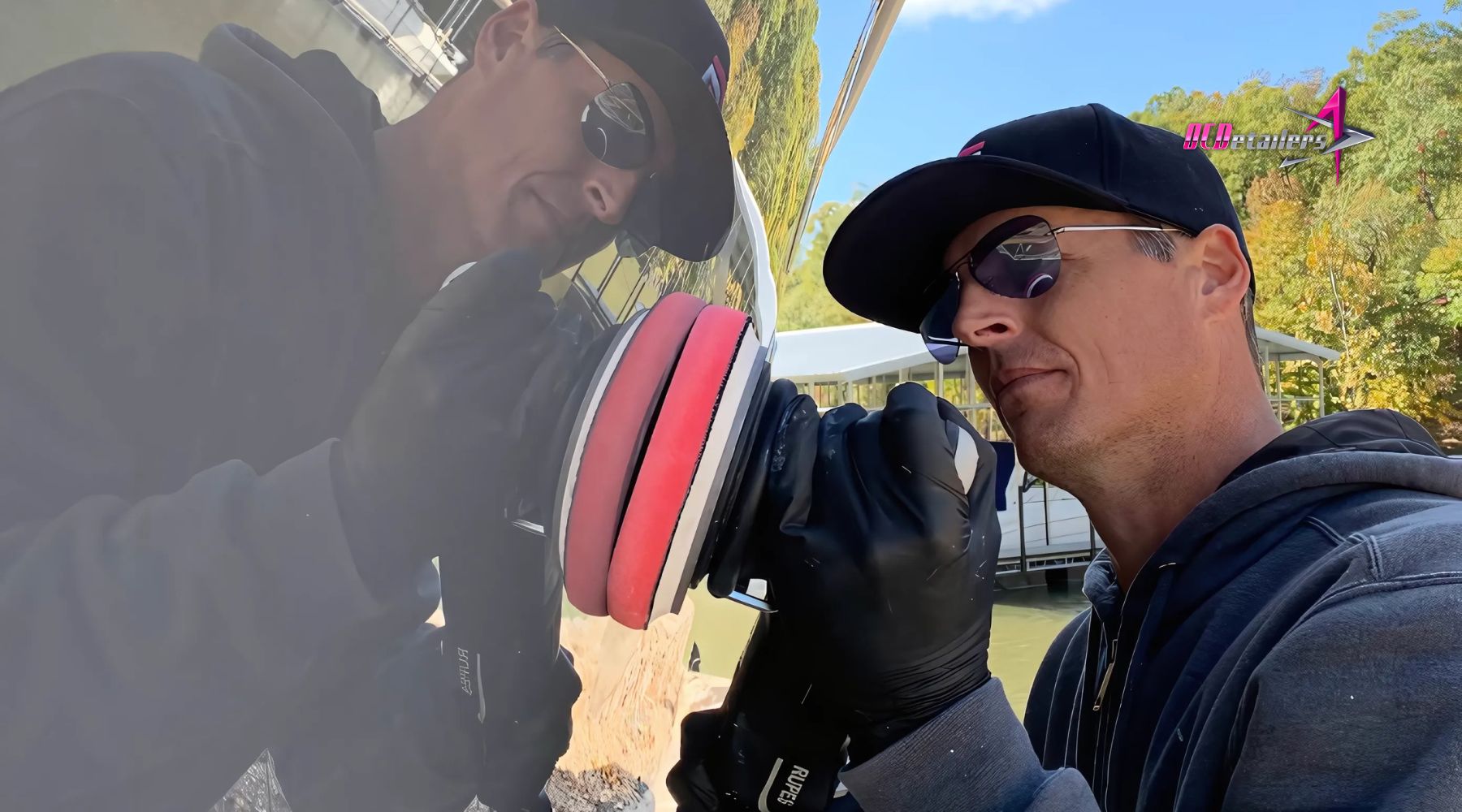 A man wearing sunglasses and a black cap uses a dual-action polisher with a red foam pad to apply marine ceramic coating to the side of a boat, reflected in the glossy surface. He’s wearing black gloves and working outdoors near a dock with trees in the background.
