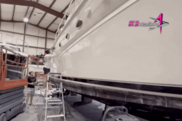 Two people work on detailing a large white yacht inside a spacious garage; one person uses a ladder to reach higher areas of the hull. The scene emphasizes the labor-intensive process of boat maintenance, with the logo "HD Detailing" visible in the top right corner.