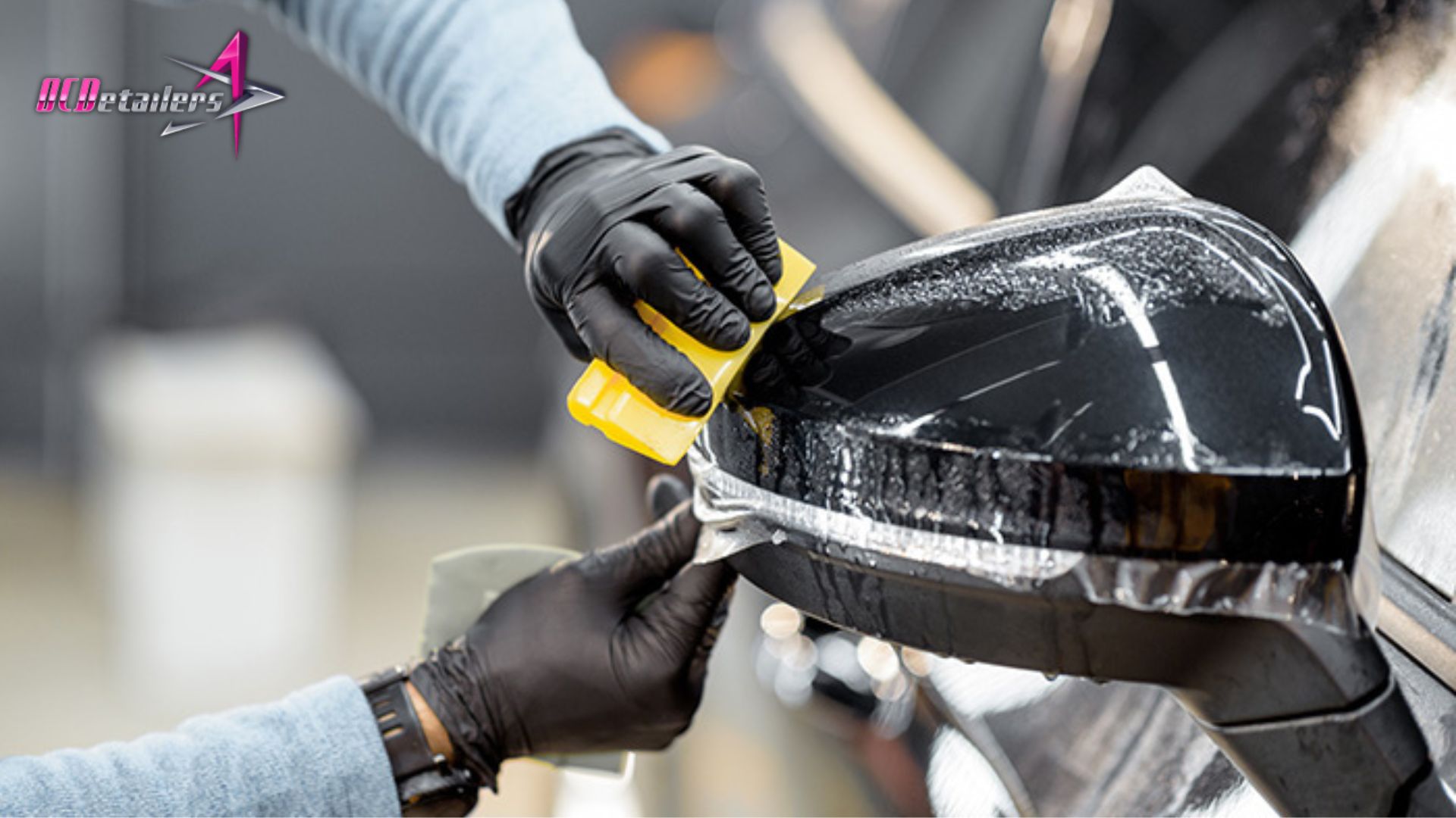 Close-up of a technician applying paint protection film to a black car side mirror using a yellow squeegee.