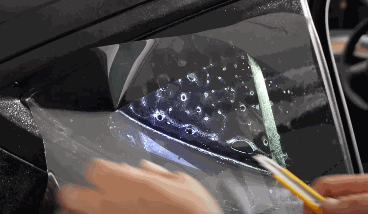Close-up of a professional applying window tinting film to a car window with a precision blade, smoothing out air bubbles for a seamless finish.