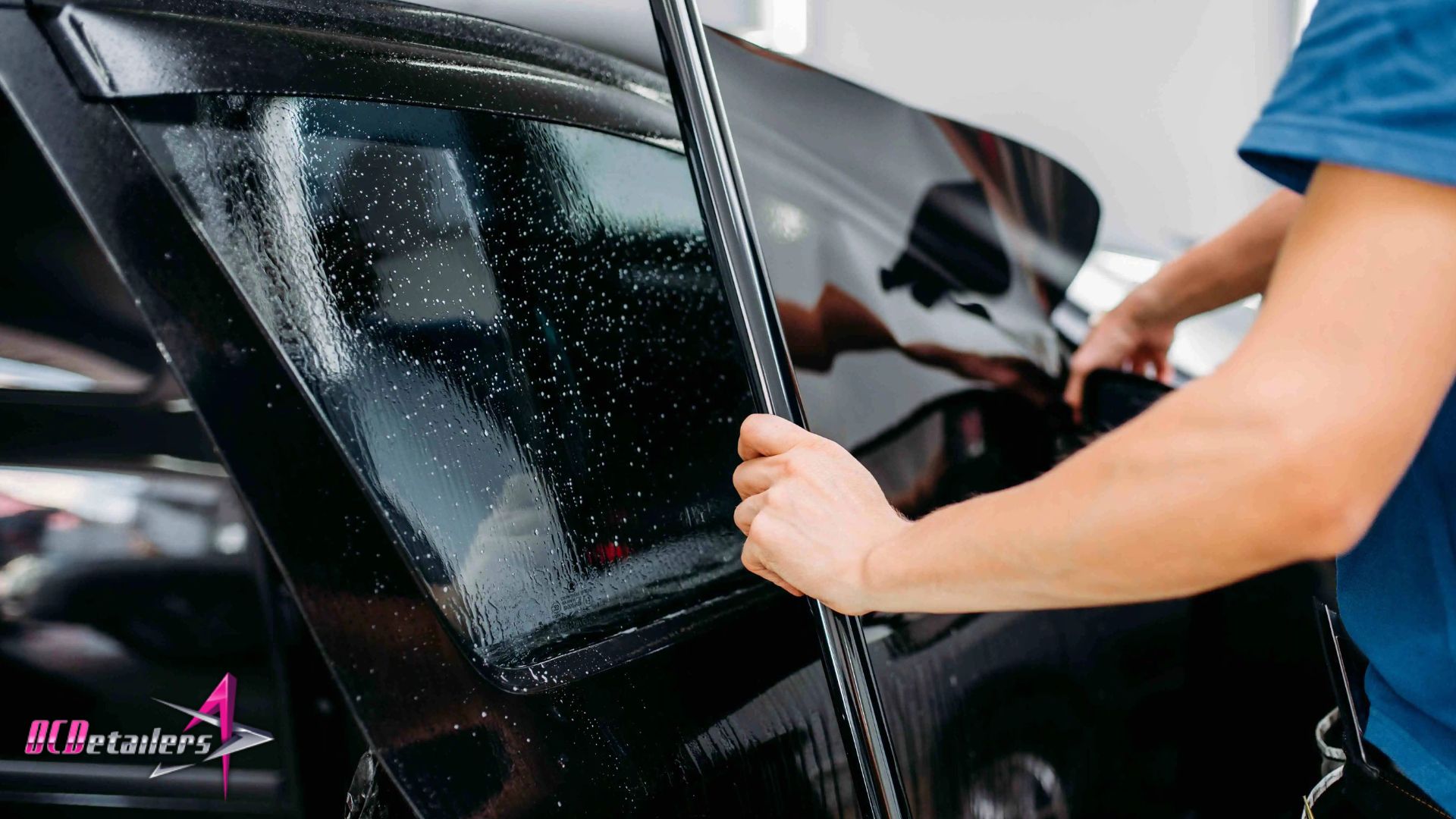 Installer holding a large sheet of window tinting film against a rear car window during the application process.