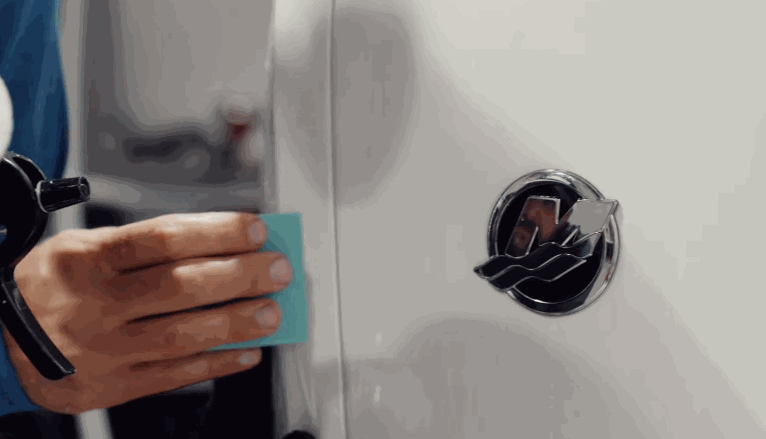 Hand pressing a blue applicator tool against a boat near a chrome emblem while applying a protective wrap.