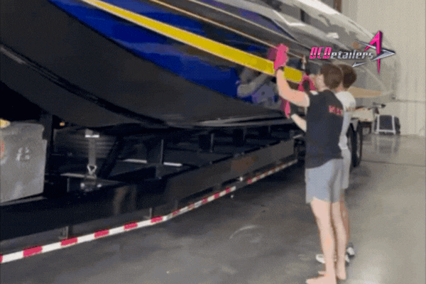Two technicians at OCDetailers inspect and polish the side of a large speedboat with bright blue and yellow detailing, using pink microfiber towels inside a garage. The boat is positioned on a trailer for easy access.