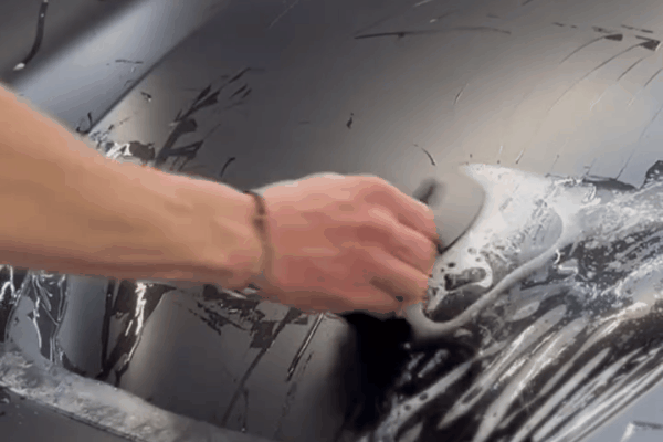 A professional applying paint protection film to a black car hood, carefully smoothing the film by hand.