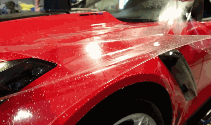 Transparent paint protection film being carefully applied to the wet surface of a red sports car's front fender.