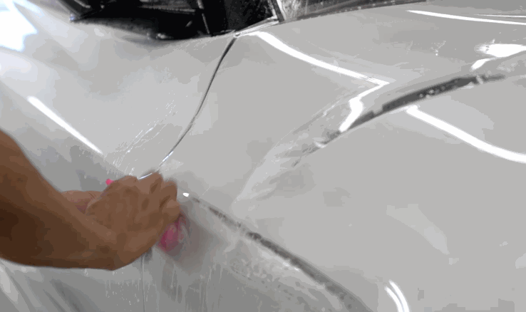Installer smoothing paint protection film over the glossy surface of a white car, using a squeegee tool on a wet panel.