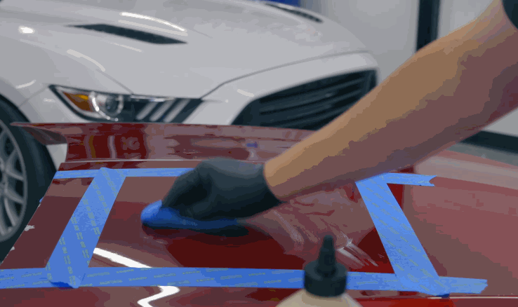 A gloved hand spreads ceramic coating on a red car panel masked with blue tape, with a white Mustang visible in the background.