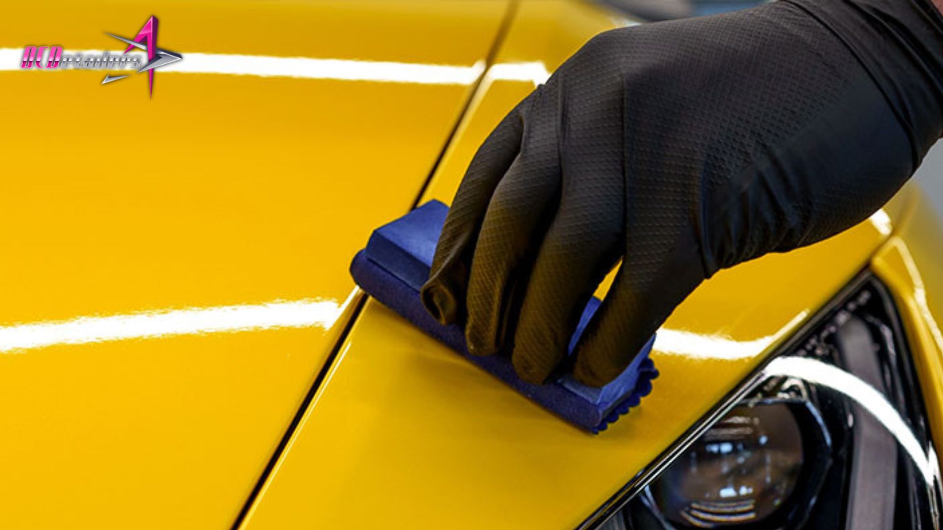 A person wearing black gloves applies ceramic coating with a blue applicator pad to the glossy yellow hood of a car.