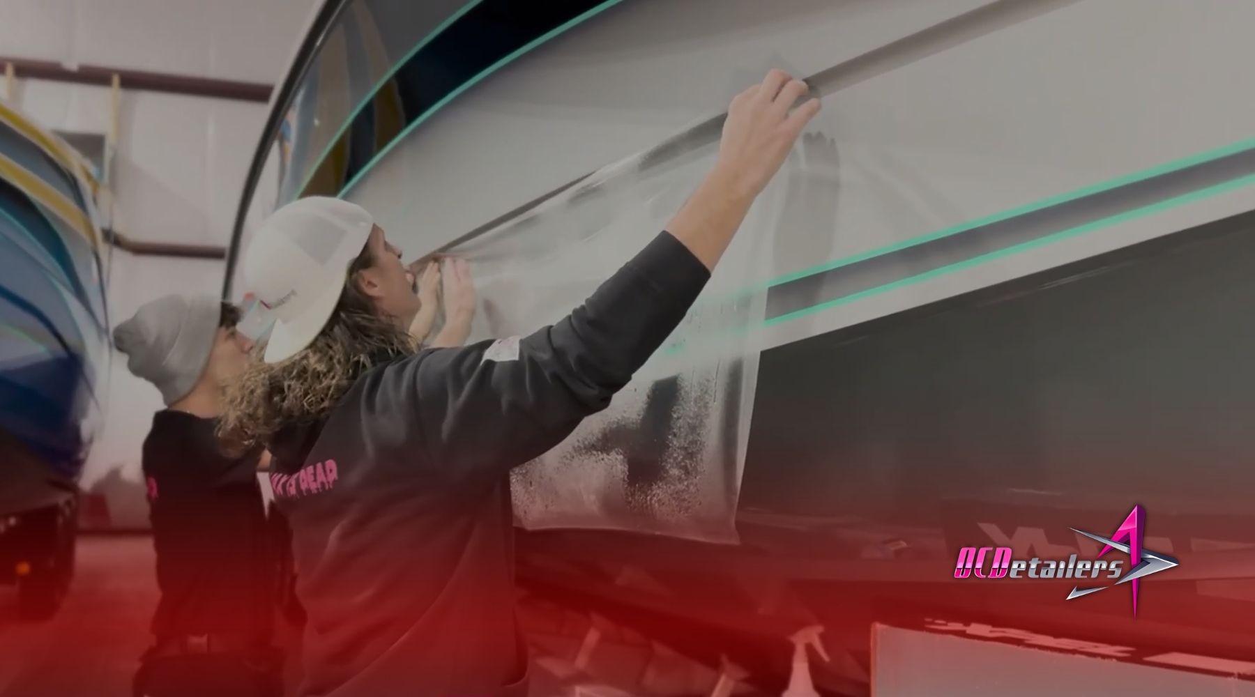 Two marine detailing professionals apply a sheet of Paint Protection Film (PPF) to the hull of a sleek boat inside a shop in Osage Beach. The process highlights the role of PPF in protecting boats from local water and weather damage common to Lake of the Ozarks.