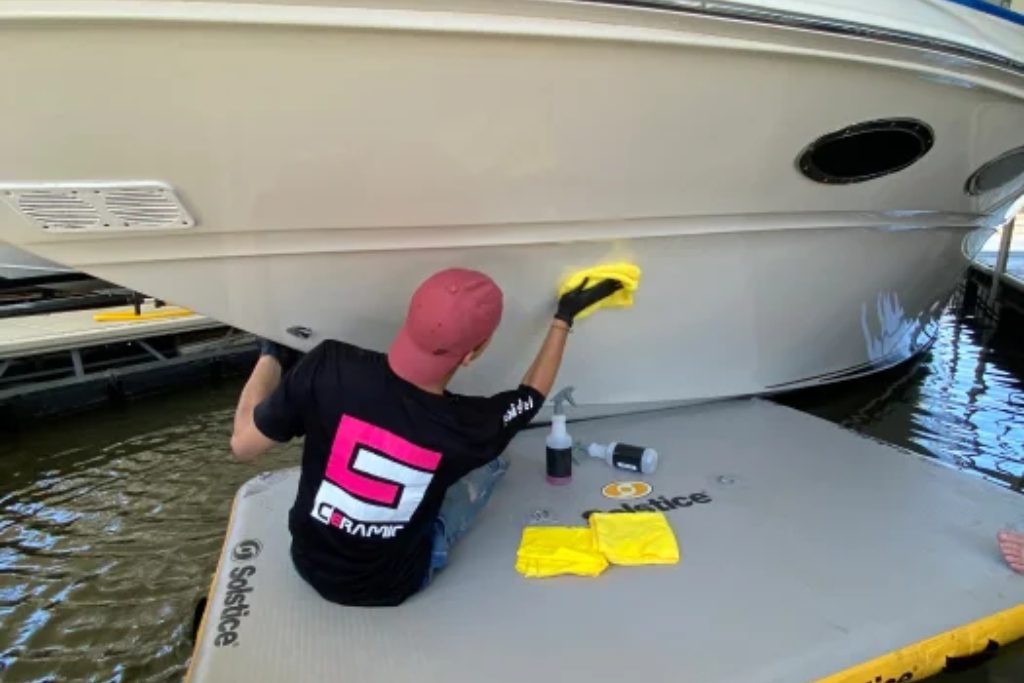 A man in a red cap and black "Ceramic" t-shirt applies a ceramic coating booster to the side of a white boat using a yellow microfiber cloth. He sits on a floating platform with spray bottles and towels beside him.